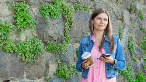 Pretty girl in a cotton jacket using smartphone with earphones and drinking Stock Footage 77065463