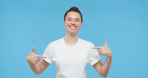 Pretty girl pointing to white t-shirt, showing empty space, isolated on blue Stock Footage
