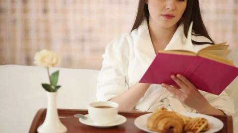 Pretty girl sitting in bed drinking tea reading book and smiling at camera Stock Footage 30949088