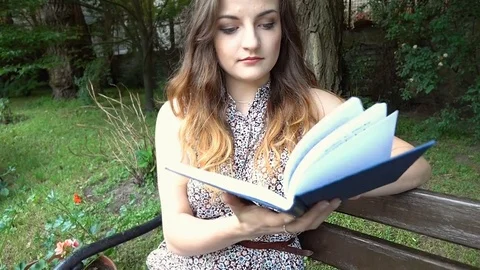 Pretty girl sitting in the park and starts reading book, steadycam shot Stock Footage