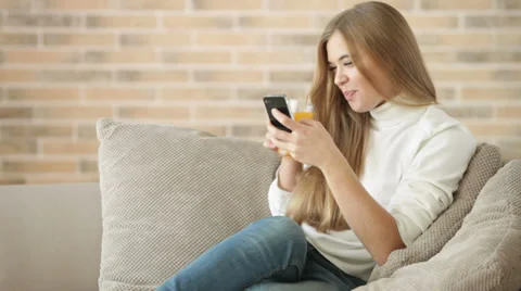 Pretty girl sitting on sofa using mobile phone holding glass of juice  Video stock 34114227
