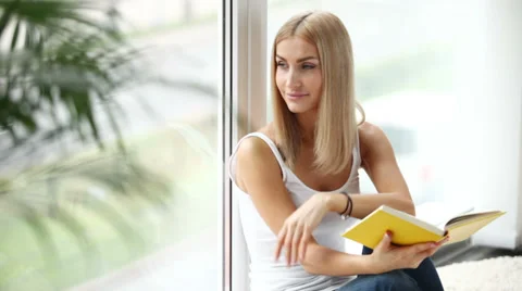 Pretty girl sitting by window reading book looking at camera and smiling Stock Footage 30554228