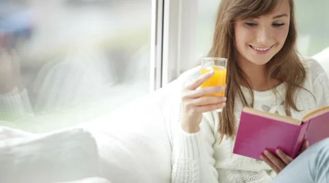 Pretty girl sitting by window reading book drinking juice looking at camera  Stock Footage 34149989