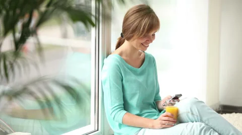 Pretty girl sitting by window using cellphone drinking juice looking at camera Vídeos de archivo 33851132