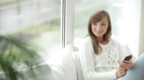 Pretty girl sitting by window using mobile phone looking at camera and smiling Stock Footage 34150039