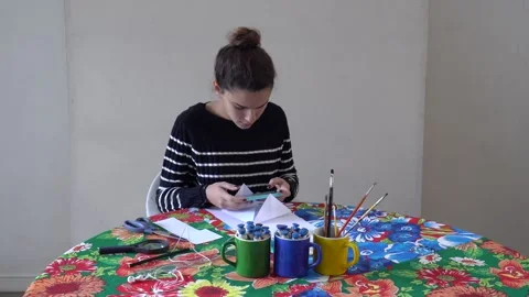 Pretty girl watches tutorial on cell phone to make origami card at art studio Stock Footage 132291853