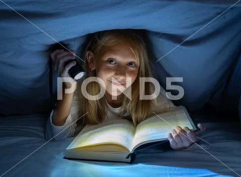 Pretty happy girl reading a book in darkness hiding under the duvet in bed wi Stock Photos