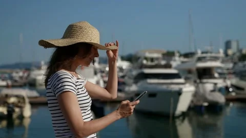 A pretty lady with a big straw hat walking along the sea with yachts uses her Video stock 158276967