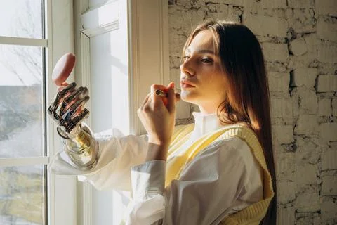 Pretty lady with bionic arm applies makeup near large window Stock Photos