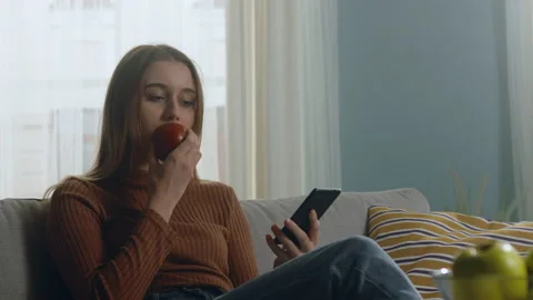 Pretty lady bites red apple and looking at something on phone 스톡 동영상 149965096