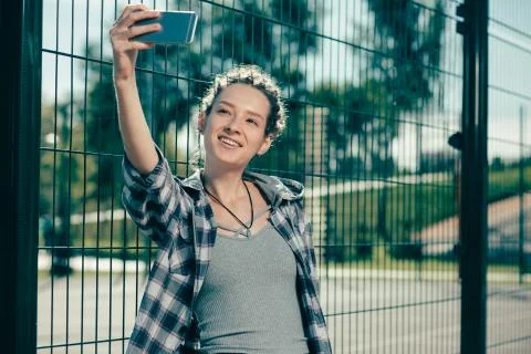 Pretty lady smiling while taking selfies near the chain link fence Stock Photos