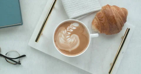 Pretty latte leaf design in white cup next to butter croissant and reading glass Stock Footage 199456832