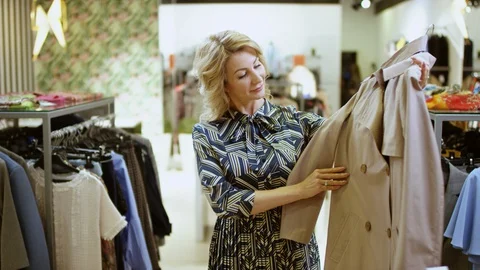 Pretty middle aged happy blond woman is trying on a coat at clothing store at Stock Footage 127921080