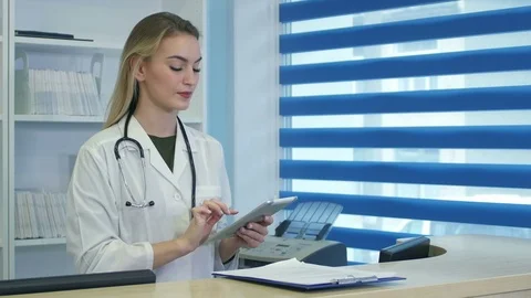 Pretty nurse using tablet and phone at hospital reception desk 스톡 동영상 77364469