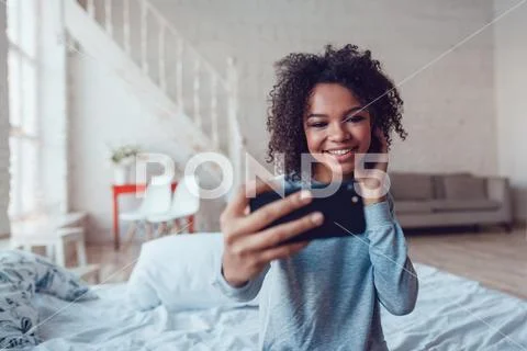 Pretty smiling african girl make selfie sitting on bed at home Stock Photos