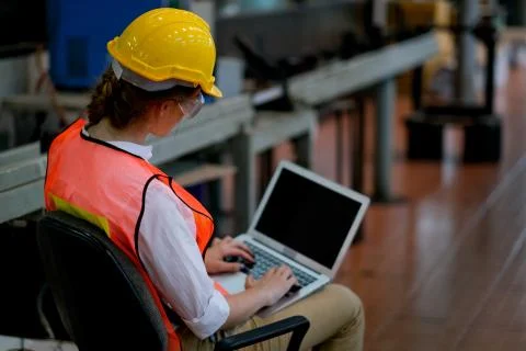 Pretty technician or engineer or worker sit on chair use laptop in factory Stock Photos