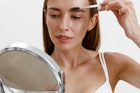 Pretty woman combing her eyebrows with brush and looking at mirror over white Stock Photos