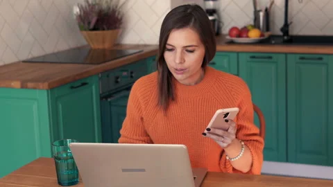 Pretty woman comparing data on smartphone and laptop in in kitchen, 4K Stock Footage 119071199