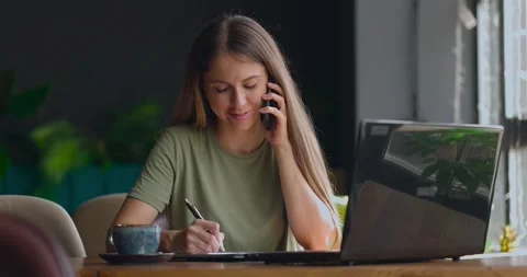 Pretty woman making notes in a notebook while speaking on phone and smiling in Video stock 159770317