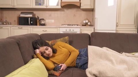 A pretty young brunette is sleeping on the corner sofa and holding a remote Stock Footage 101388766