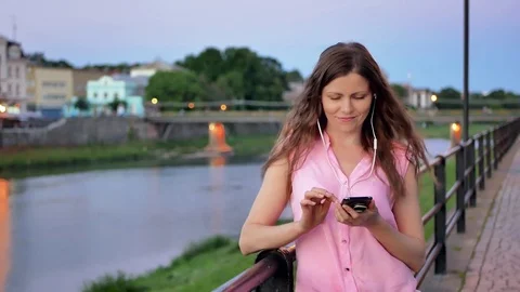 Pretty young girl using smartphone with earphones near the railing on waterfront Stock Footage 77148756