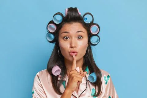 Pretty young woman in big hair curlers wearing silk top presses index finger to Stock Photos