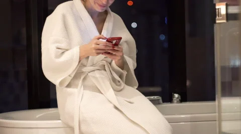 Pretty, young woman sitting in bathrobe with smartphone in bathroom Stock Footage 59125166