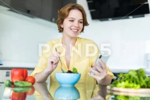 Pretty young woman using her mobile phone while eating salad in the kitchen a Stock Photos