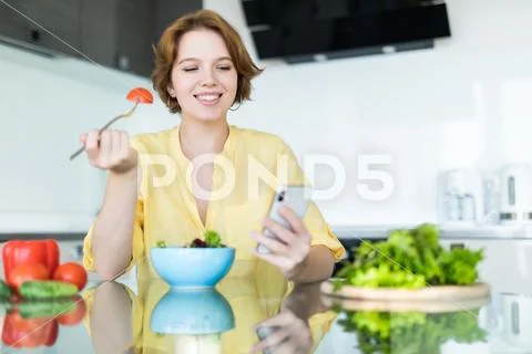 Pretty young woman using her mobile phone while eating salad in the kitchen a Stock Photos