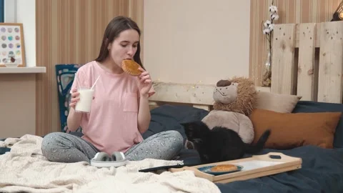 Prety yound girl in payjamas eats cookie, drinks milk, in bed near with cat Stock Footage 167511429