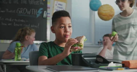 Primary School Boy Sitting at the Desk, Eating Sandwich from Lunch Box, Talking Stock Footage 314022210