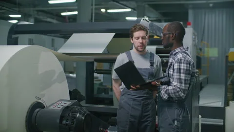 Printing Factory Operators Analyzing Data on Laptop near Large Paper Reel Stock Footage 335483798