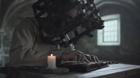 Prisoner in an iron head cage running a finger along text in an open book 스톡 동영상 339230865