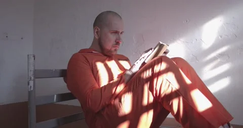 Prisoner in orange overalls reading a book in a prison cell sitting on the bed. Vídeos de archivo 167765626