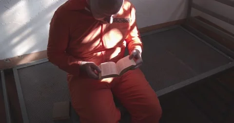 Prisoner in orange uniform reading a book in a prison cell. 스톡 동영상 232333955