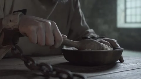 Prisoner in shackles lined with fabric eating porridge inside a gloomy cell Stock Footage 340322726