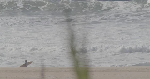 Pro surfer walking on sandy beach next to ocean with big waves splashing Stock Footage 160766081