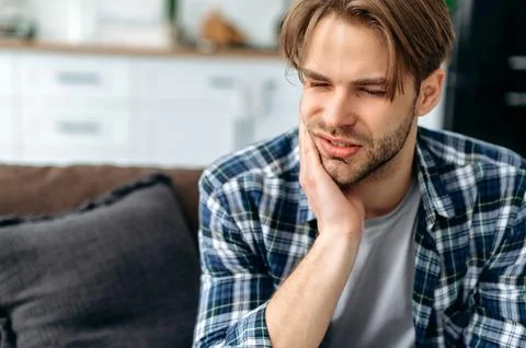 Problems with teeth. Toothache. Upset caucasian guy is sitting on the couch at Stock Photos