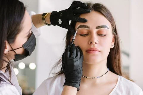 The process of dyeing the eyebrows of a young brunette by an eyebrow master with Stock Photos