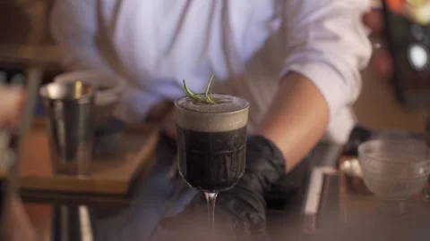 Professional Bartender Serving Dark Craft Cocktail with Rosemary Garnish Stock Footage 330976753