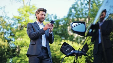 Professional Businessman Uses Smartphone While Charging Electric Car Outdoors Stock-Footage 312865530