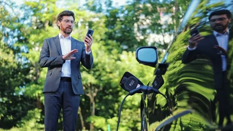 Professional Businessman on Video Call While Charging His Electric Car Outdoors Video stock 312865645
