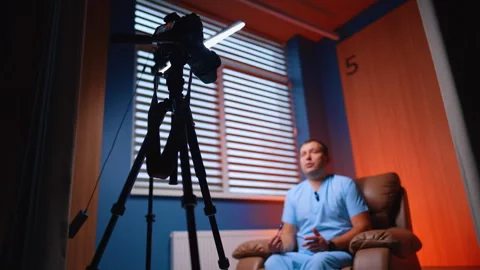 Professional camera and soffit are arranged in front of the man in armchair. Low Stock Footage 334339303