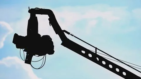 Professional cinema camera on a crane arm, silhouetted against a pale blue sky Stock Photos