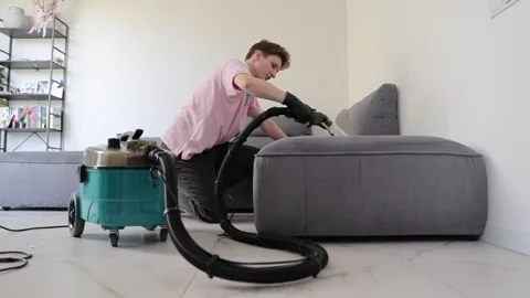 Professional cleaner using vacuum to refresh gray sofa in a bright stylish Stock Footage 321515448