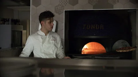 Professional focused pizza chef taking a pizza out of the oven and preparing to Stock Footage 199457914