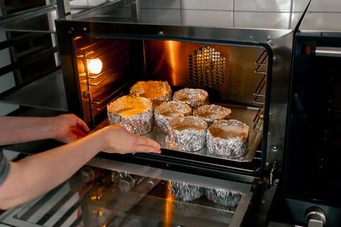 In a professional kitchen a baker inserts a baking tray with cake molds into the Stock Photos