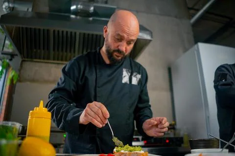 In a professional kitchen, a chef in a black jacket puts ingredients on toast Stock Photos