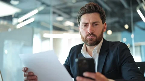 Professional Man Deeply Focused Comparing Business Documents with Smartphone Stock Footage 329274731