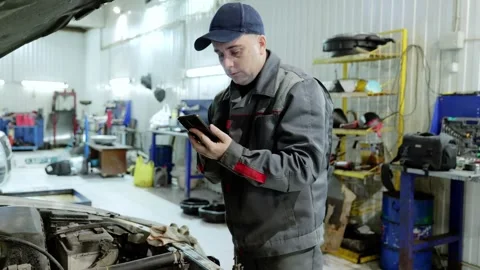 Professional mechanic checking smartphone while inspecting car engine, using Stock Footage 341289575